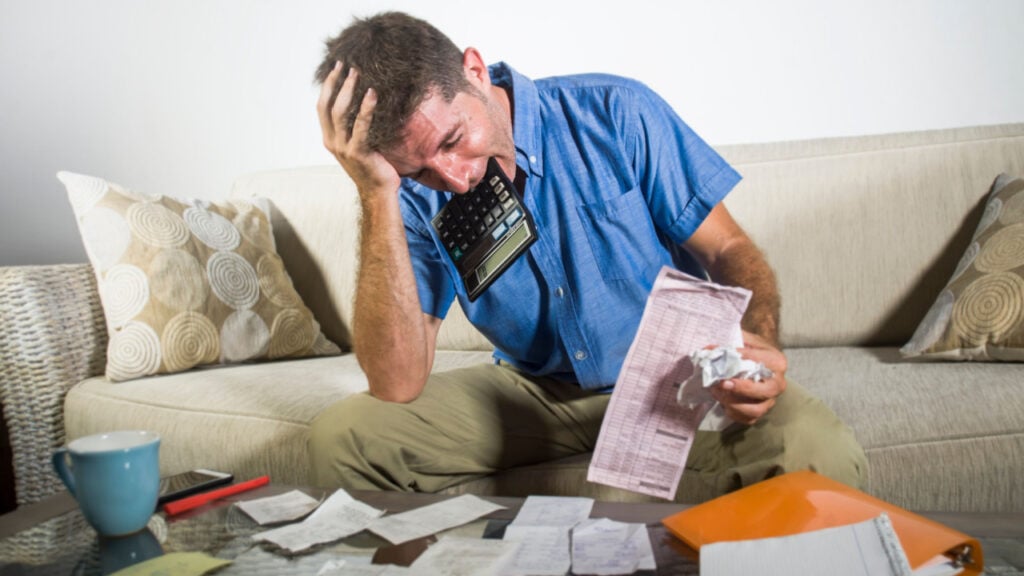 young stressed and overwhelmed man biting calculator holding mess of bank and receipts paperwork desperate calculating monthly expenses taxes and income frustrated in domestic accounting