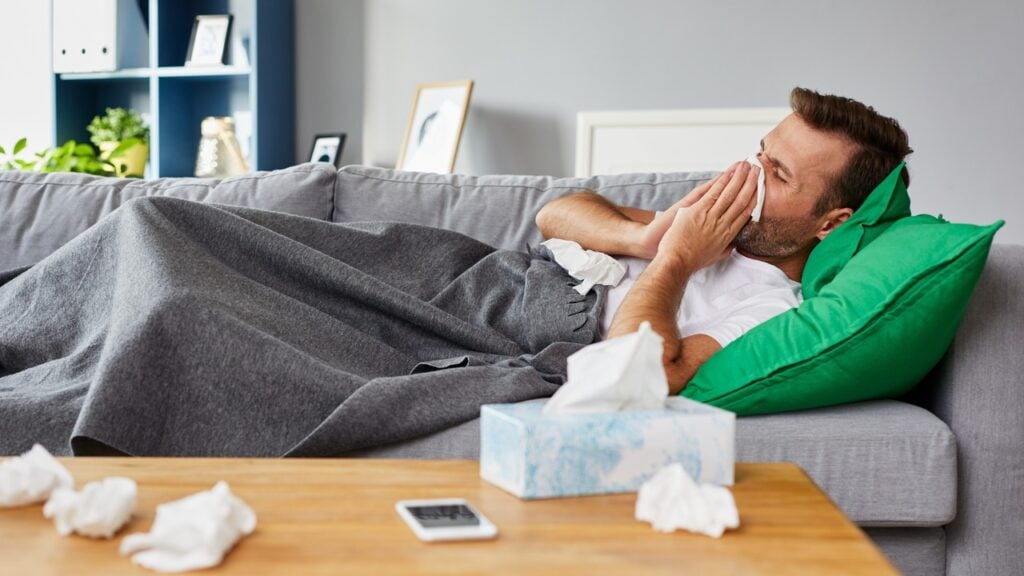 A man sick on the couch at home, blowing his nose into kleenex.