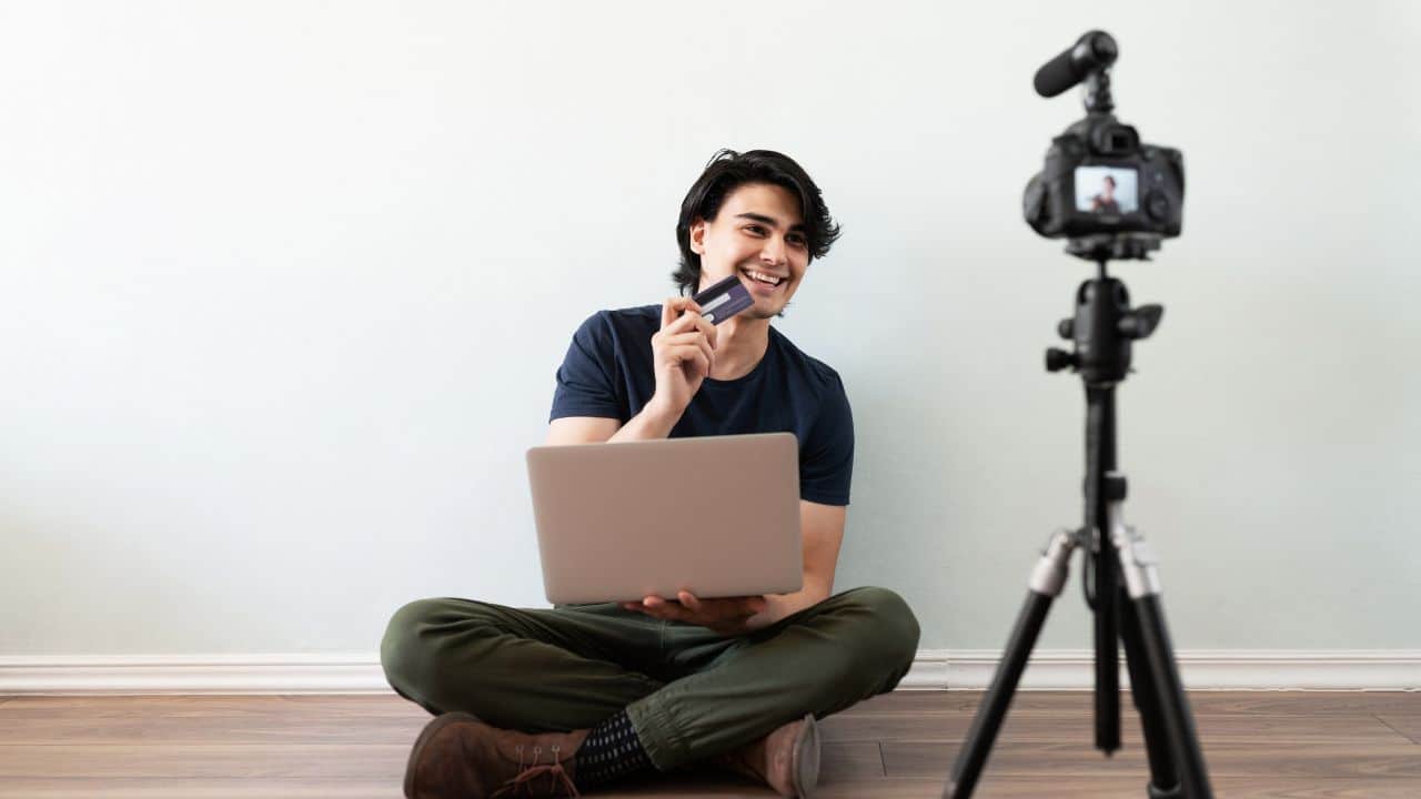 Man, blogger filming himself with a camera, holding a laptop, holding a credit card, wearing a black shirt