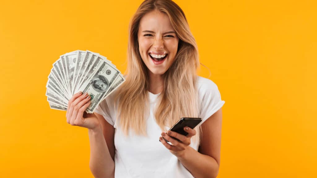 Smiling blonde woman holding phone and cash money yellow background