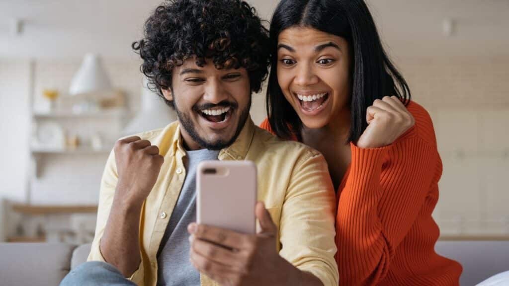 Excited friends using smartphone playing mobile game relaxing at home. Happy emotional couple win online lottery celebration success. Multiracial students reading test results